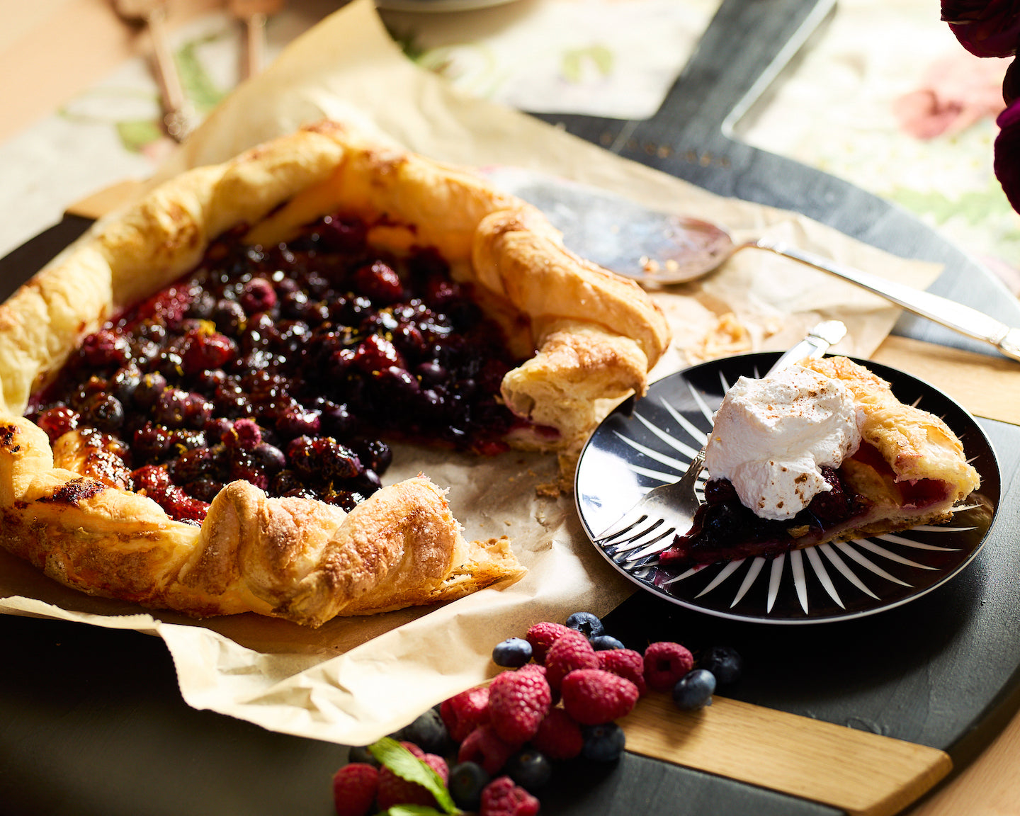 A blueberry crostata on a sheet of parchment paper next to a small Marrakech plate with a slice of the dessert topped with whipped cream.
