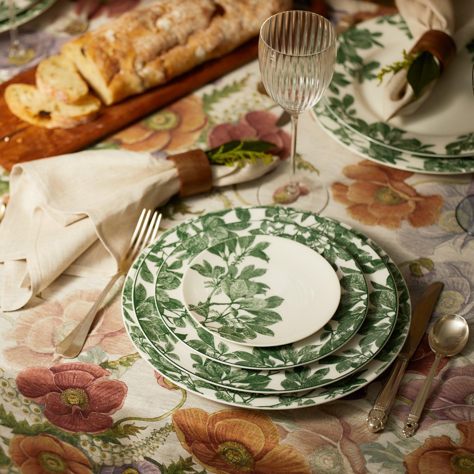 A table setting with plates featuring green foliage patterns, silverware, a glass, a napkin with a wooden ring, and a loaf of bread on a floral tablecloth.