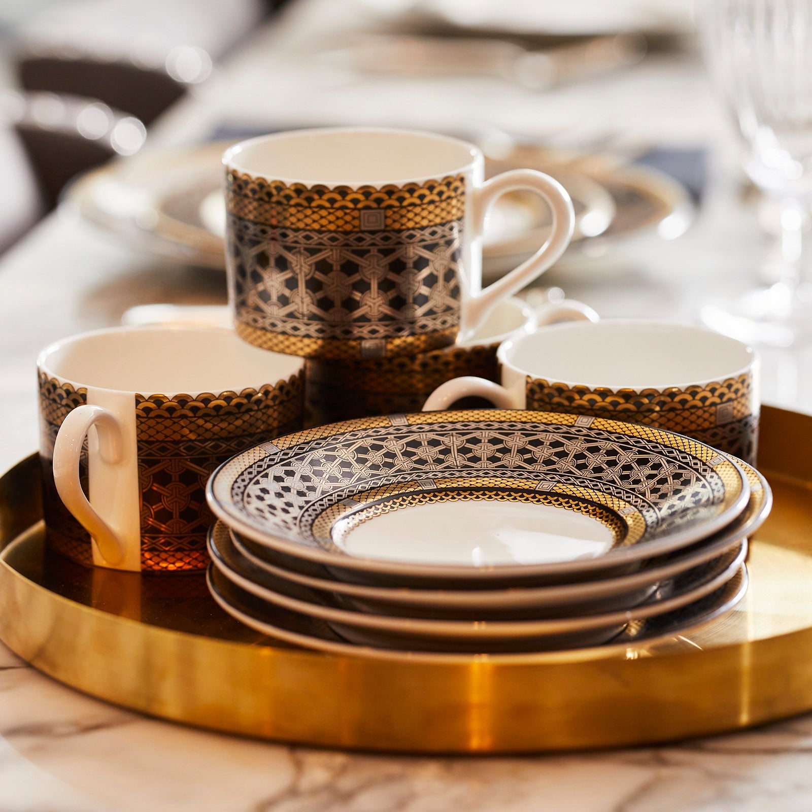 A set of decorative plates and coffee mugs with intricate designs sits on a round gold tray placed on a marble table.