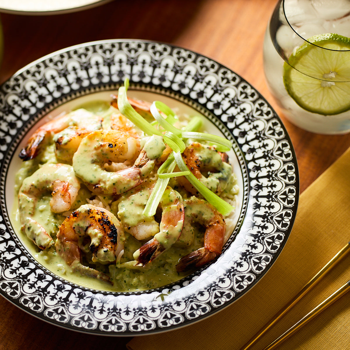 A plate of shrimp in a creamy green sauce garnished with sliced green onions, served on beautiful Casablanca Rimmed Soup Bowl by Caskata Artisanal Home with hand decorated details, accompanied by a drink garnished with a lime slice next to it.