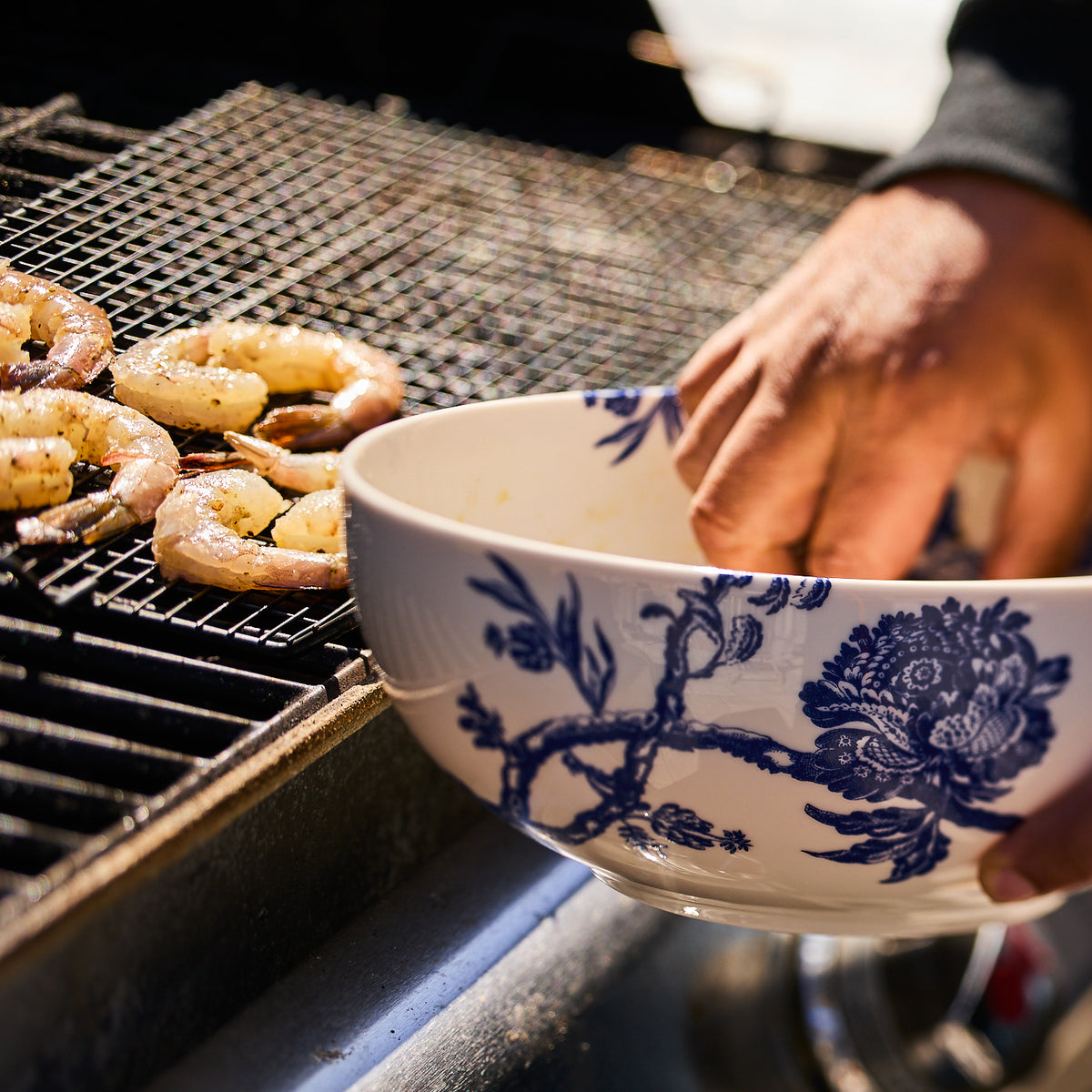 A person is placing seasoned shrimp on a grill using an Arcadia Vegetable Serving Bowl, reminiscent of the elegance found in Caskata Artisanal Home&#39;s premium porcelain dinnerware.