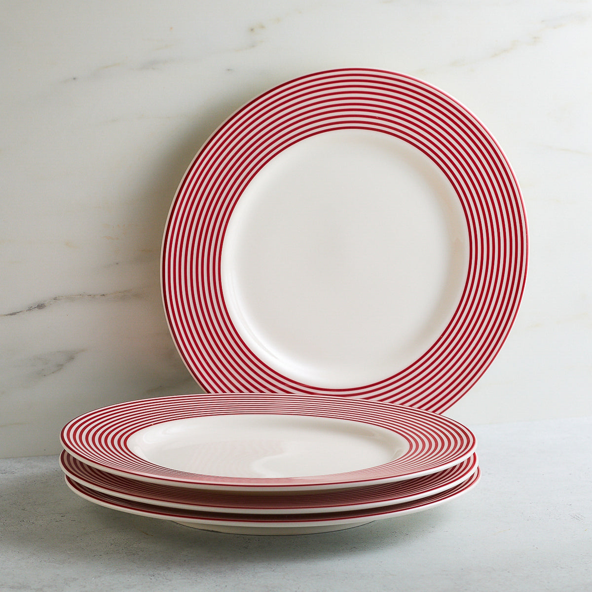 A stack of four Caskata Artisanal Home Newport Stripe Crimson Dinner Plates, showcasing white surfaces adorned with red concentric rings, is placed on a light-colored surface. An additional plate with the same design and casual elegance is propped upright behind the stack.