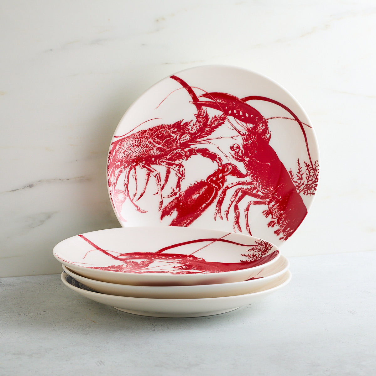 A stack of round white ceramic plates with red lobster illustrations, placed on a light-colored surface against a white marble background, perfectly matches the charming July Fourth Bundle by Caskata hanging nearby.