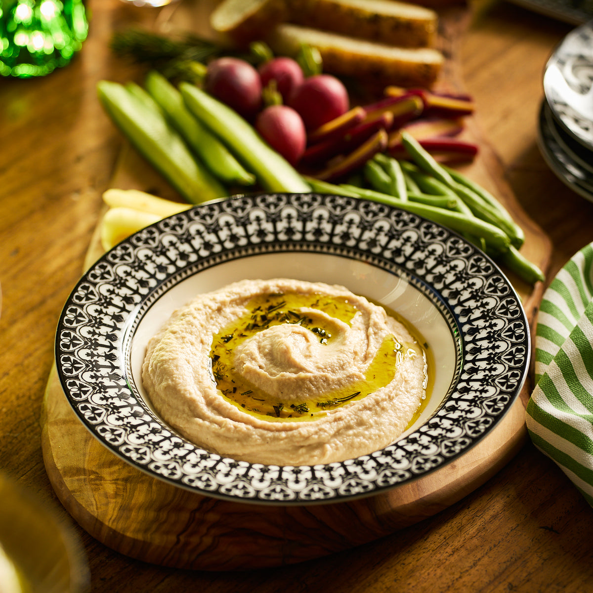 A bowl of hummus garnished with olive oil and herbs, presented in a classic Casablanca Rimmed Soup Bowl by Caskata Artisanal Home, surrounded by sliced bread, cucumber, radishes, green beans, and bell pepper on a wooden board.