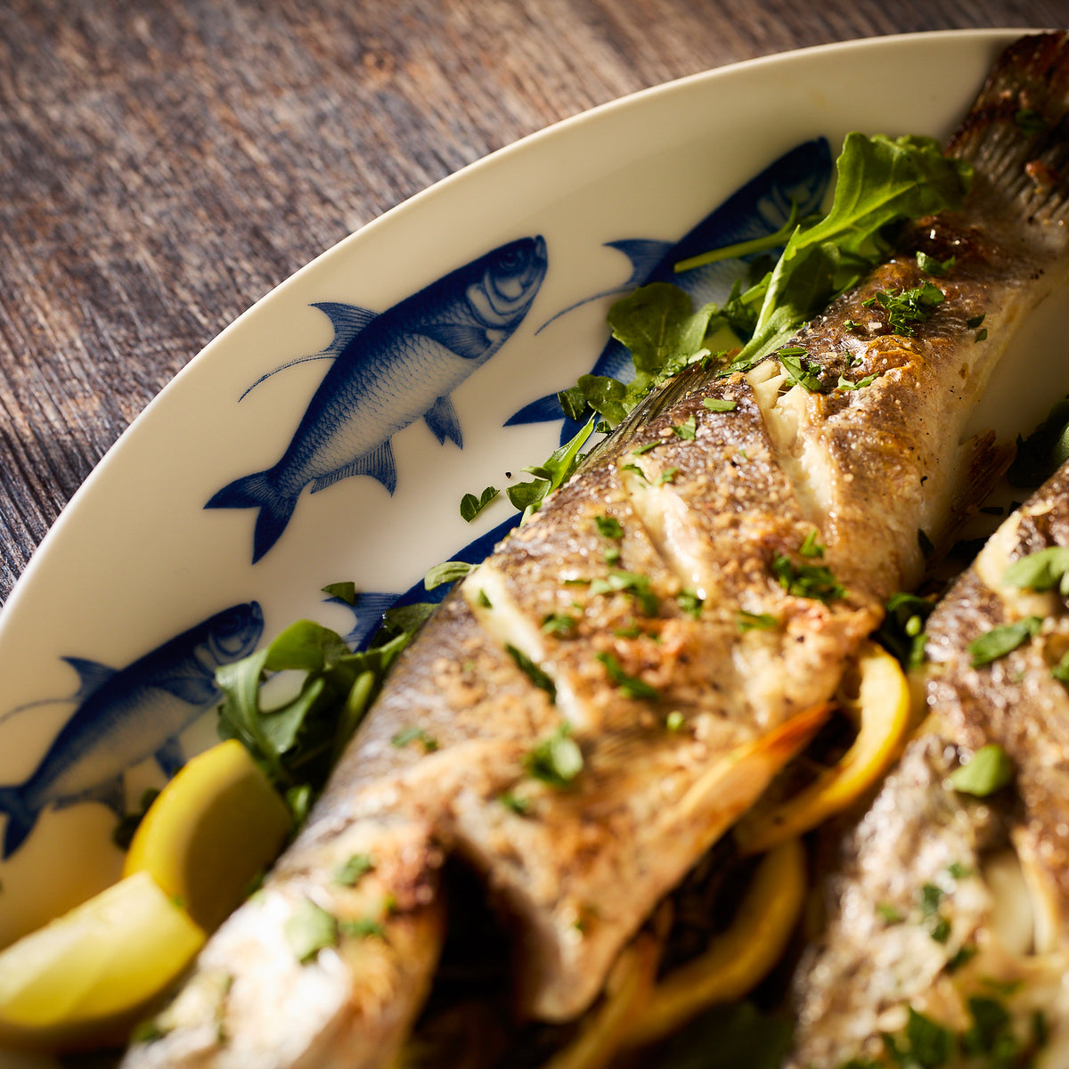 A cooked fish garnished with herbs and lemon slices is served on a white plate with fish illustrations, reminiscent of a cozy lake house meal, placed on a wooden surface, using the Lake House Bundle by Caskata.