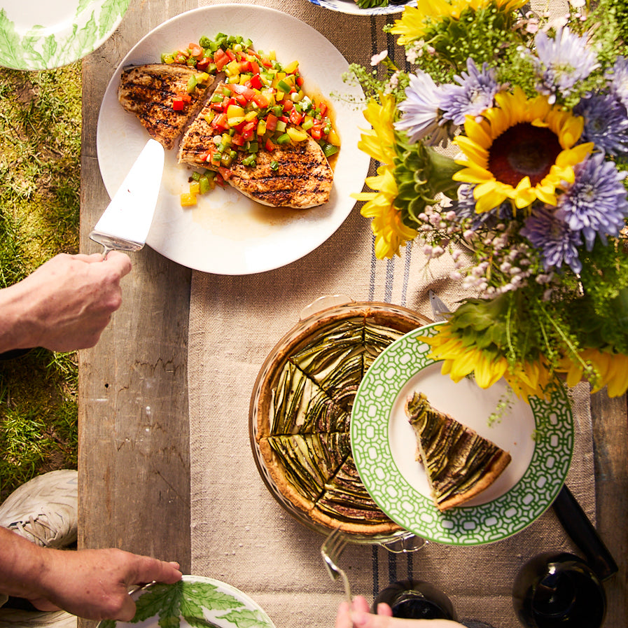 A table set with grilled chicken topped with diced vegetables, a quiche-like dish, and Newport Garden Gate Verde Rimmed Salad Plates by Caskata Artisanal Home made of premium porcelain. A vase of sunflowers and other flowers adds a contemporary note, as hands reach for the food.