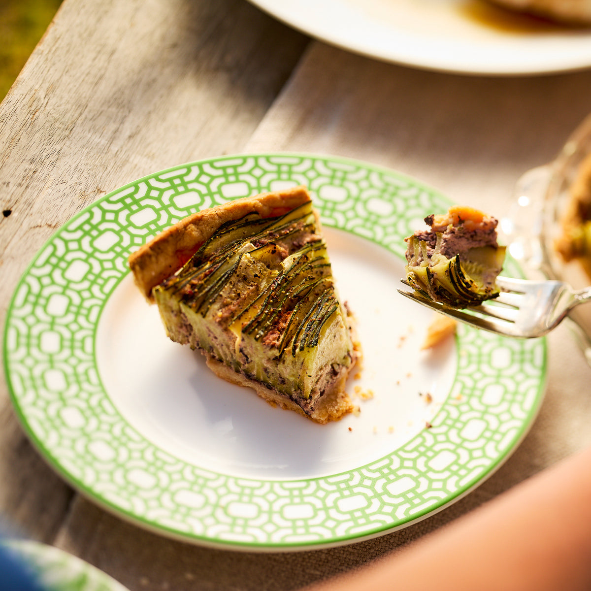 A slice of vegetable quiche is on a green patterned plate from Caskata&#39;s Through the Garden Gate Dinner Bundle, with a fork holding a small piece.