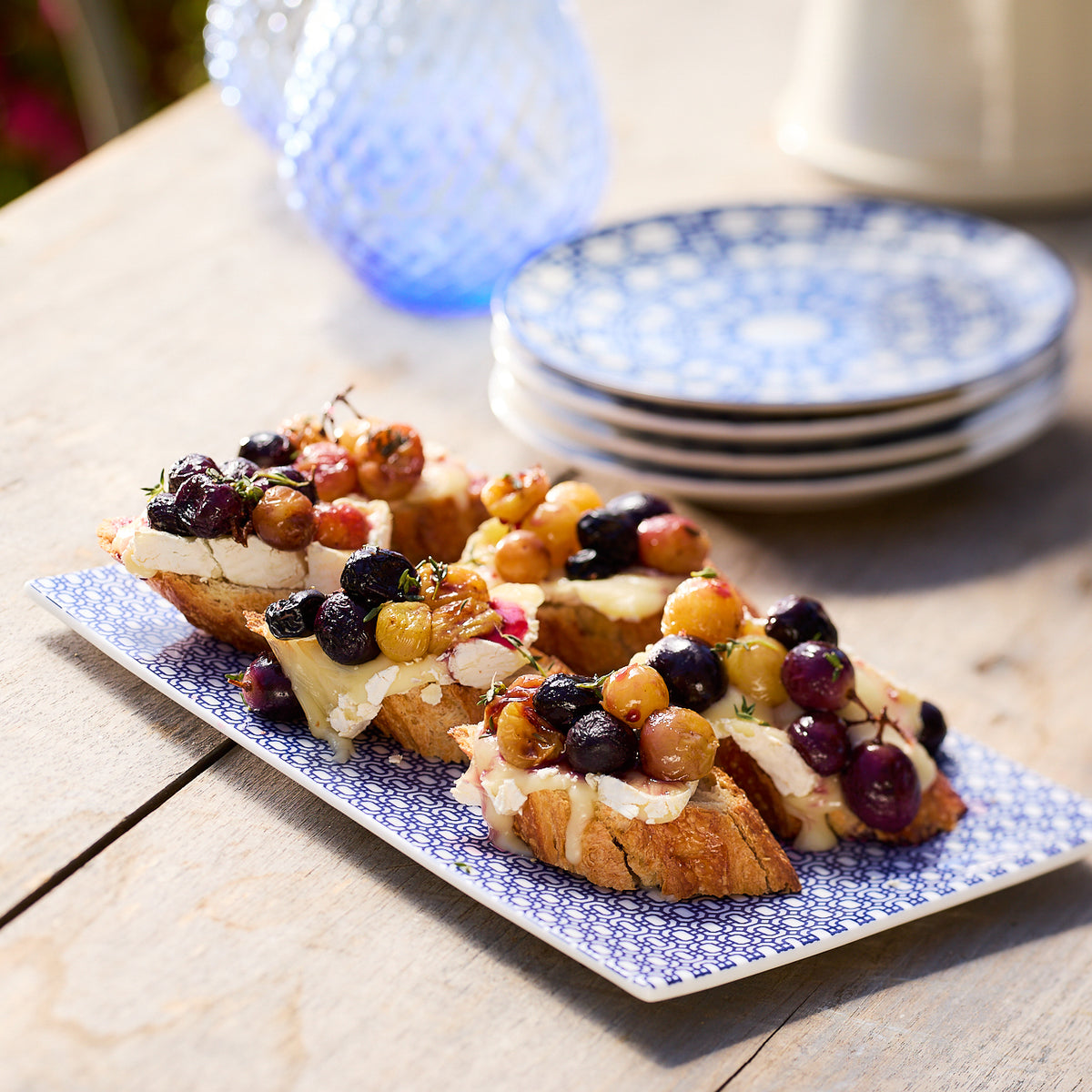 Slices of toasted bread topped with various berries and cream cheese are arranged on a Caskata Newport Large Sushi Tray, set on a wooden table next to a stack of premium porcelain plates and a blue glass jug.