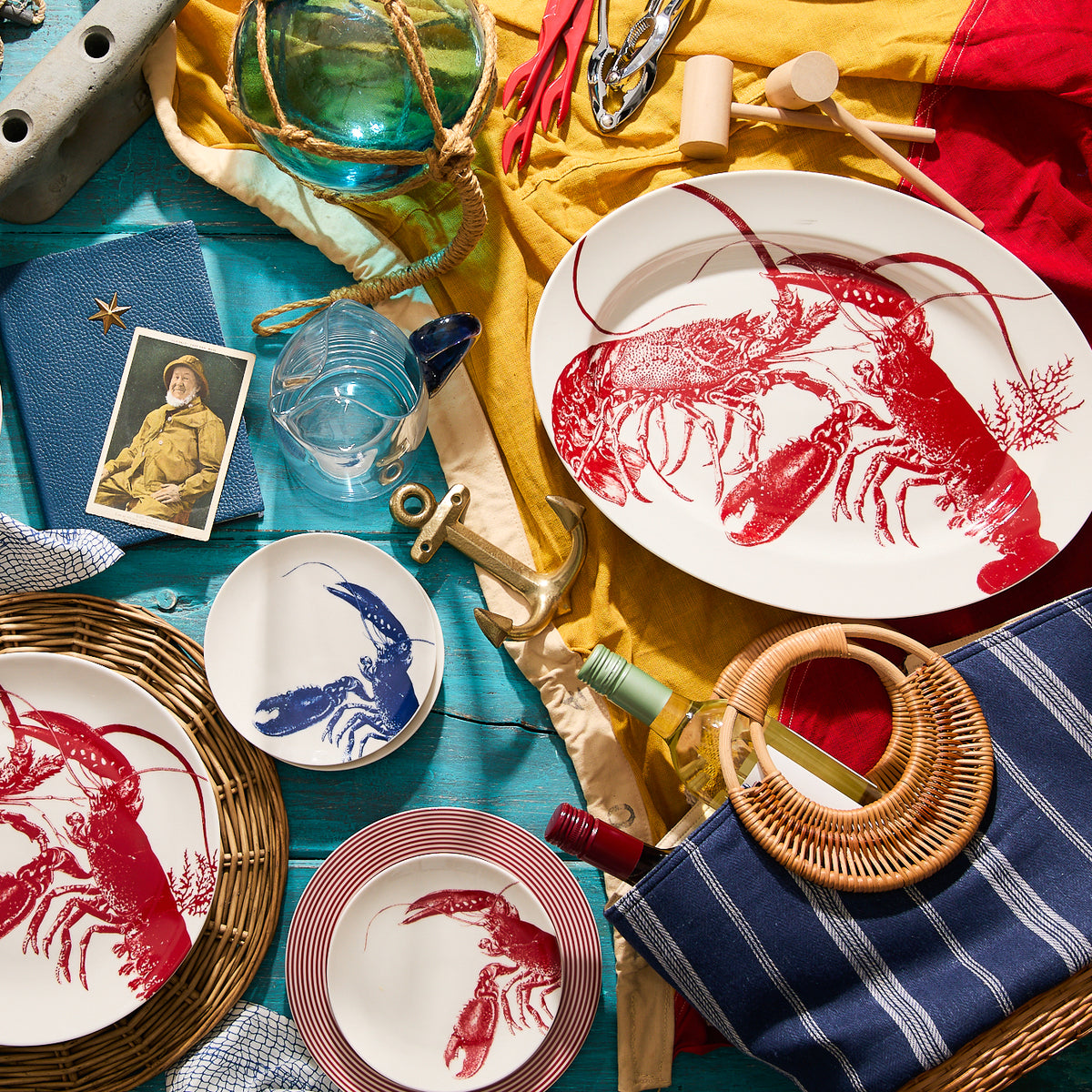 A Caskata Artisanal Home Lobster Oval Rimmed Platter and various nautical items, including an old photo, folded yellow and red fabric, glass floats, and a striped cloth, arranged on a blue surface in true seaside style.