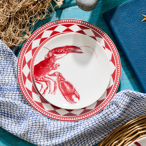 A white Fez Crimson Rimmed Salad Plate from Caskata Artisanal Home with a red lobster illustration is placed on a larger red and white patterned plate. The plates are on a blue surface with a fish net, blue cloth, and a woven basket nearby, evoking the charm of Moroccan patterns.