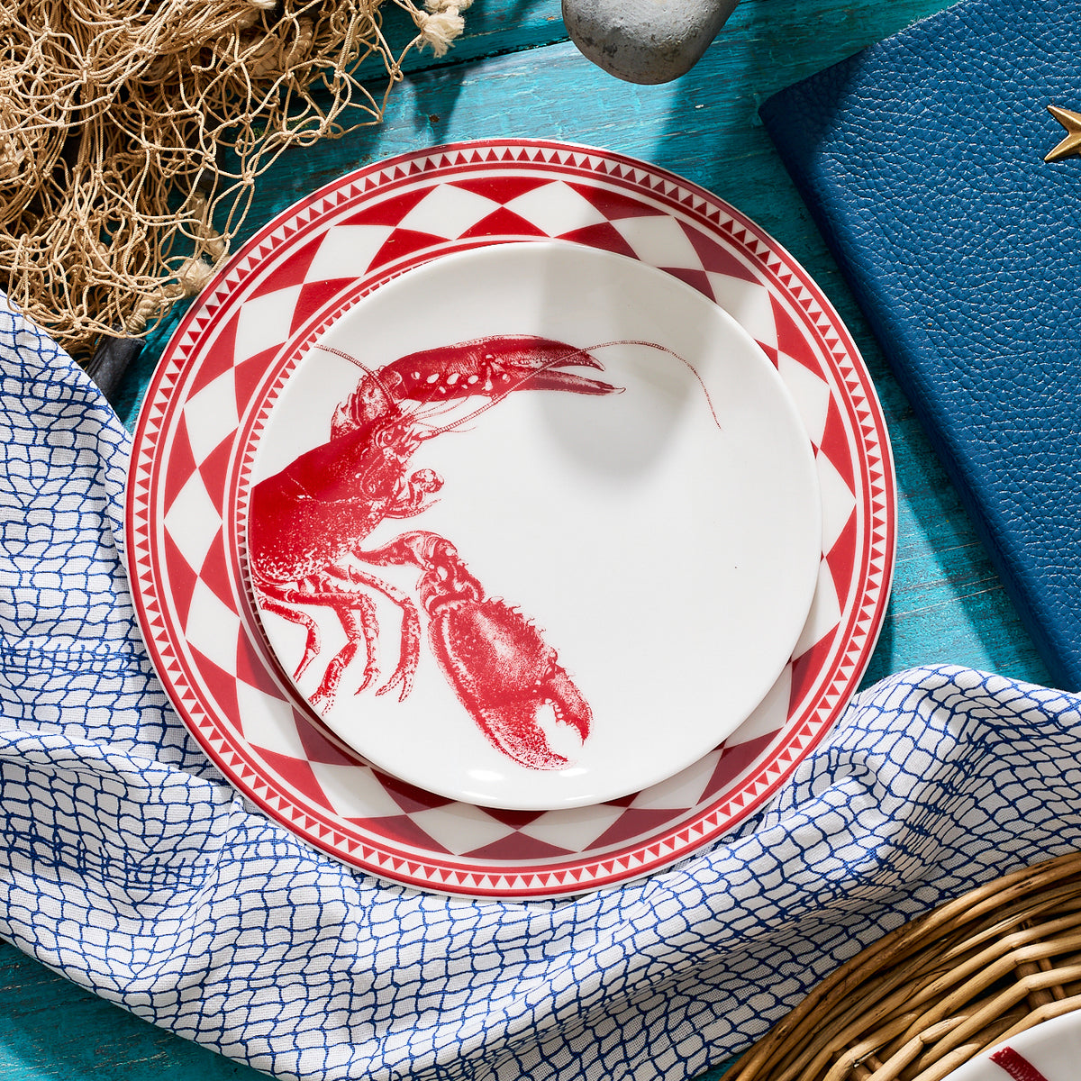 A white plate with a red lobster design, part of the Caskata Artisanal Home heirloom-quality Lobster Red Small Plates dinnerware, sits atop a larger red and white geometric-patterned plate on a blue table adorned with a white and blue cloth, a woven basket, netting, and a blue book reminiscent of the New England coast.