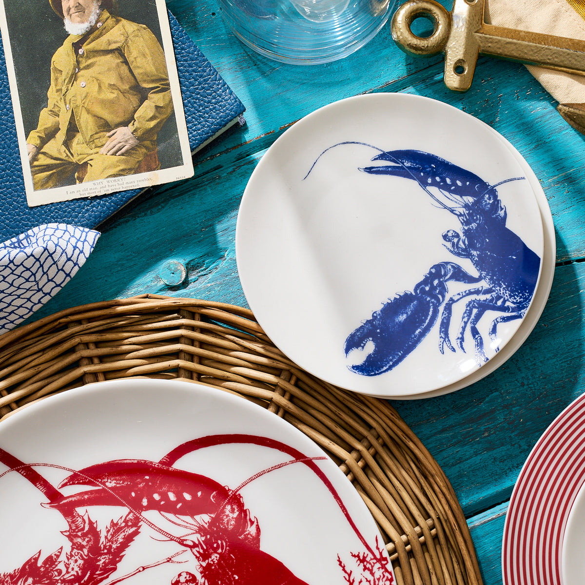 Two heirloom-quality &quot;Lobster Small Plates&quot; by Caskata Artisanal Home, one with a red lobster and the other with a blue lobster design, sit on a blue table with a rattan placemat and a photograph of a person in vintage clothing. This seaside style dinnerware adds a charming touch to any coastal-themed dining experience.