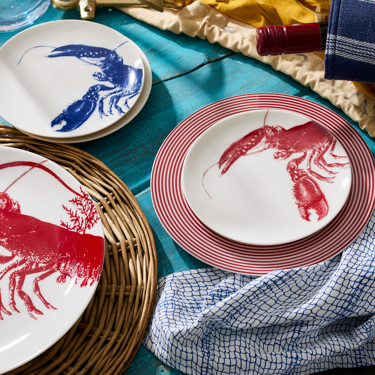 A set of heirloom-quality Lobster Red Small Plates by Caskata Artisanal Home, some with red lobsters and some with blue lobsters, arranged on a blue wooden table next to a wine bottle and a blue and white checkered cloth, evoke a charming seaside style.