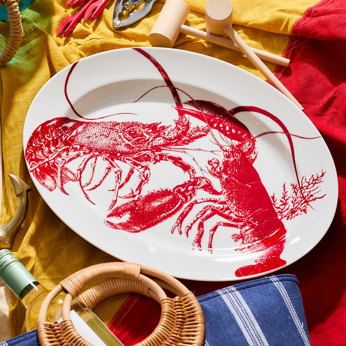 A Lobster Oval Rimmed Platter by Caskata Artisanal Home with red illustrations is surrounded by various kitchen and dining utensils, including wooden mallets, a can opener, and a blue and white striped fabric, evoking a charming seaside style.