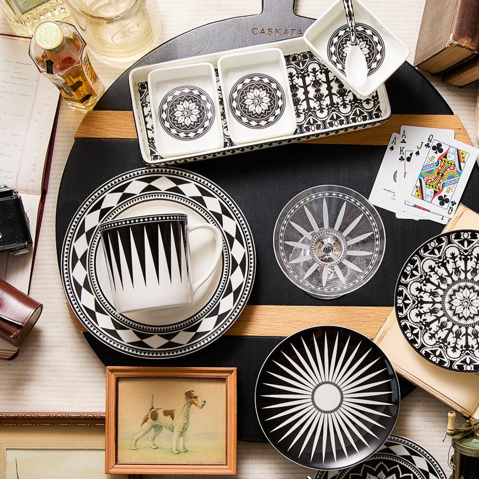 A table set with the Caskata Casablanca Nested Appetizer Tray &amp; Spoon Set, featuring black and white geometric patterns, along with a framed dog illustration, playing cards, a drink, and other various objects creates an inviting scene.