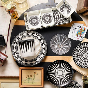 A table set with the Caskata Casablanca Nested Appetizer Tray & Spoon Set, featuring black and white geometric patterns, along with a framed dog illustration, playing cards, a drink, and other various objects creates an inviting scene.