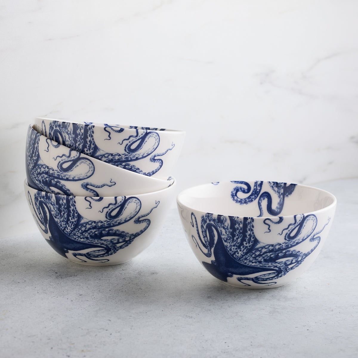 Four Lucy Cereal Bowls by Caskata made from premium porcelain with intricate blue designs. Three are stacked on the left, and one is placed to the right on a grey surface against a marble backdrop. These dishwasher safe bowls are perfect for any meal or decor.