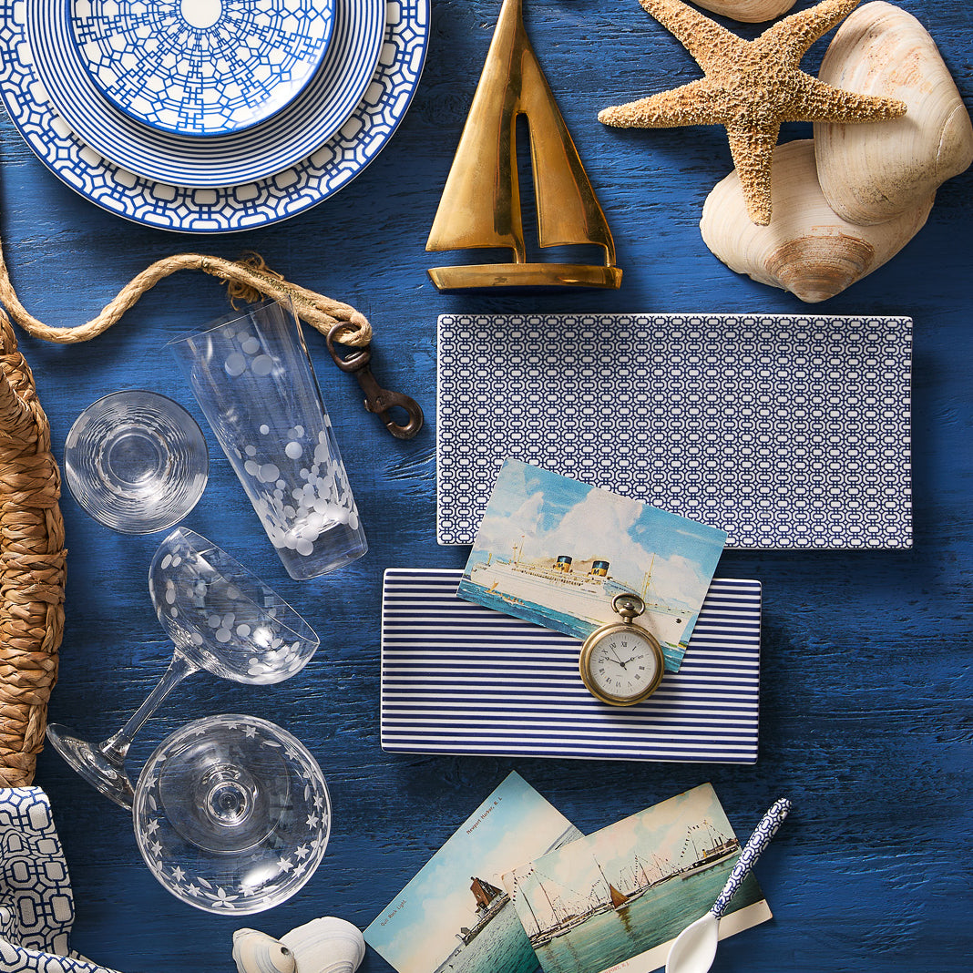 A nautical-themed flat lay featuring blue-patterned dishes, premium porcelain glassware, seashells, starfish, a brass sailboat, Newport Large Sushi Tray by Caskata postcards, a watch, and a wicker basket on a blue wooden surface.