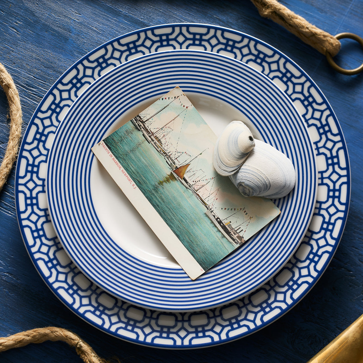 A nautical-themed table setting featuring a blue and white patterned Newport Garden Gate Rimmed Dinner Plate by Caskata Artisanal Home, a postcard with a ship illustration, and two seashells.