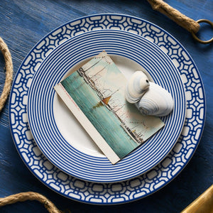 A Newport Stripe Rimmed Salad Plate from Caskata Artisanal Home holds a vintage postcard depicting boats and a seashell, placed on a blue wooden surface with two thick ropes in the background, exuding a touch of casual elegance.