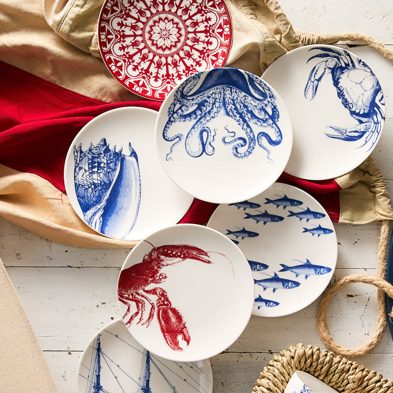 Collection of Shells Small Plates by Caskata Artisanal Home, lead-free porcelain plates with blue and red nautical designs, including octopus, crab, fish, lobster, and seashells, arranged on a textured fabric surface. This heirloom-quality dinnerware offers a whimsical touch to any meal.