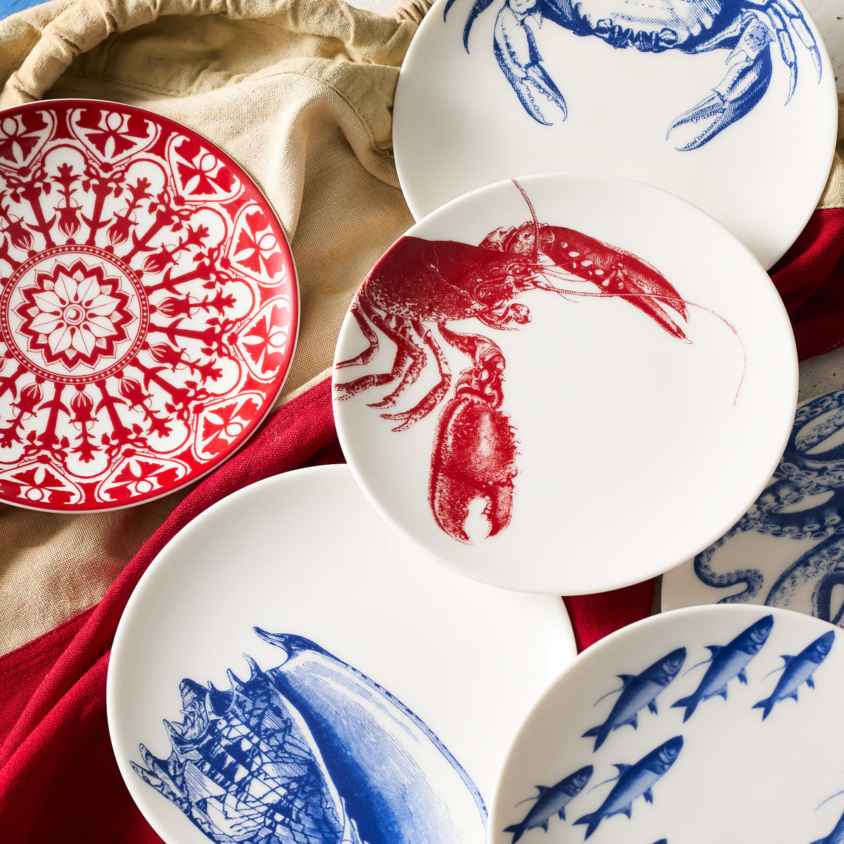 A selection of Caskata Artisanal Home&#39;s Lobster Red Small Plates, featuring white plates with red and blue designs, including intricate patterns, a lobster, a crab, and various sea creatures inspired by New England&#39;s coast, all arranged on a red and beige cloth background.