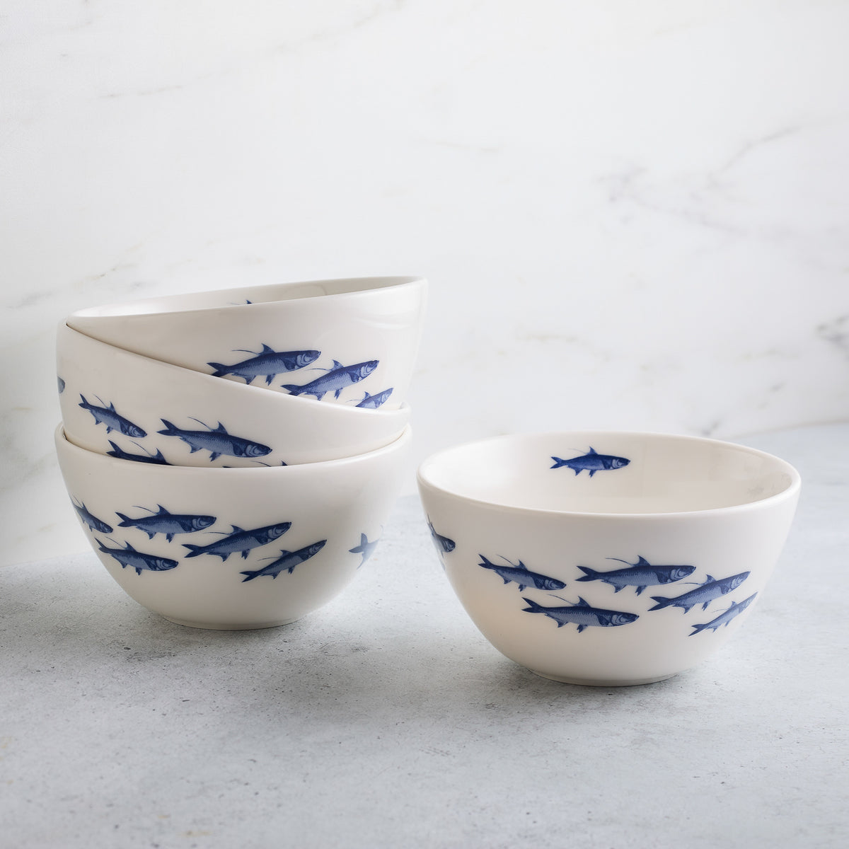 A stack of four Caskata School of Fish Cereal Bowls, each decorated with a pattern of blue fish, makes a charming addition to any dinnerware collection.