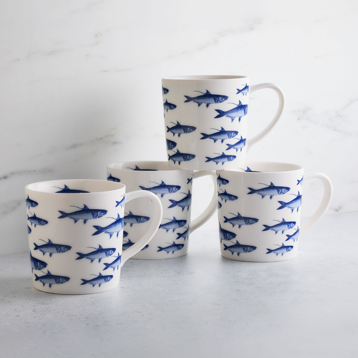 Four white ceramic mugs featuring brilliant blue fish patterns are arranged in a semi-circle on a light gray surface with a marble-like background. These high-fired porcelain School of Fish Mugs by Caskata Artisanal Home add elegance to any dinnerware collection.