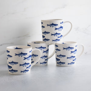 Four white ceramic mugs featuring brilliant blue fish patterns are arranged in a semi-circle on a light gray surface with a marble-like background. These high-fired porcelain School of Fish Mugs by Caskata Artisanal Home add elegance to any dinnerware collection.