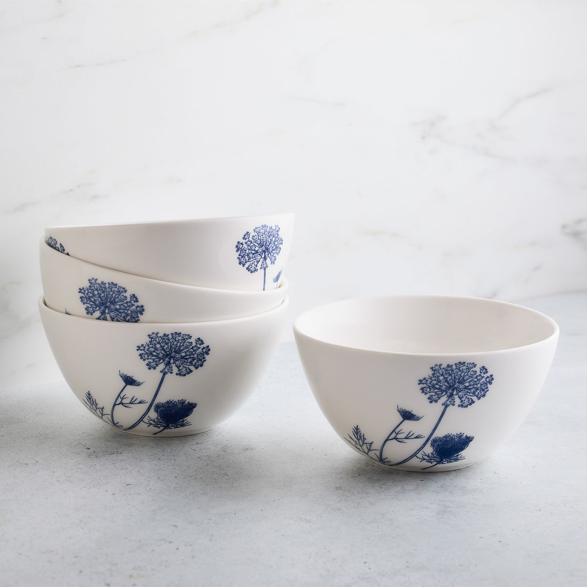 Four Caskata Summer Blues Cereal Bowls with blue floral designs, part of the Summer Blues collection, are stacked on a light gray surface against a white marble background.