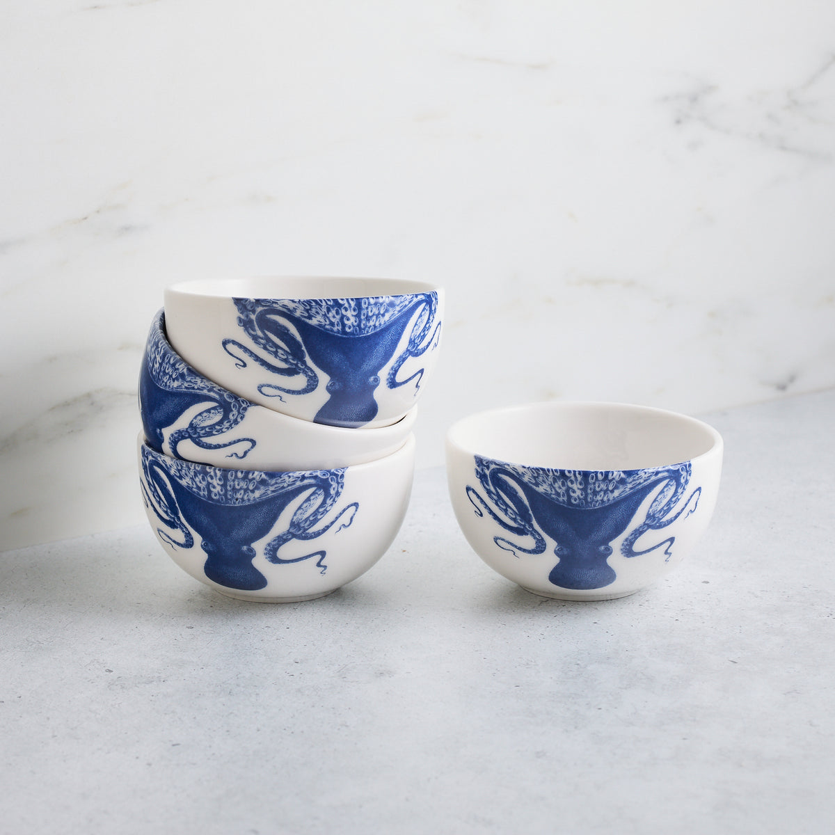 Three white Lucy Snack Bowls with blue Lucy the octopus designs by Caskata Artisanal Home are stacked, and one matching small bowl is placed beside the stack on a light gray surface, against a marble backdrop.