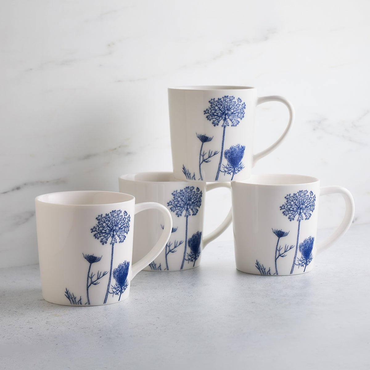 Four white ceramic mugs with blue floral designs, arranged stacked on a light surface, against a marble background. This set of Summer Blues Mugs from Caskata Artisanal Home is perfect for any occasion.