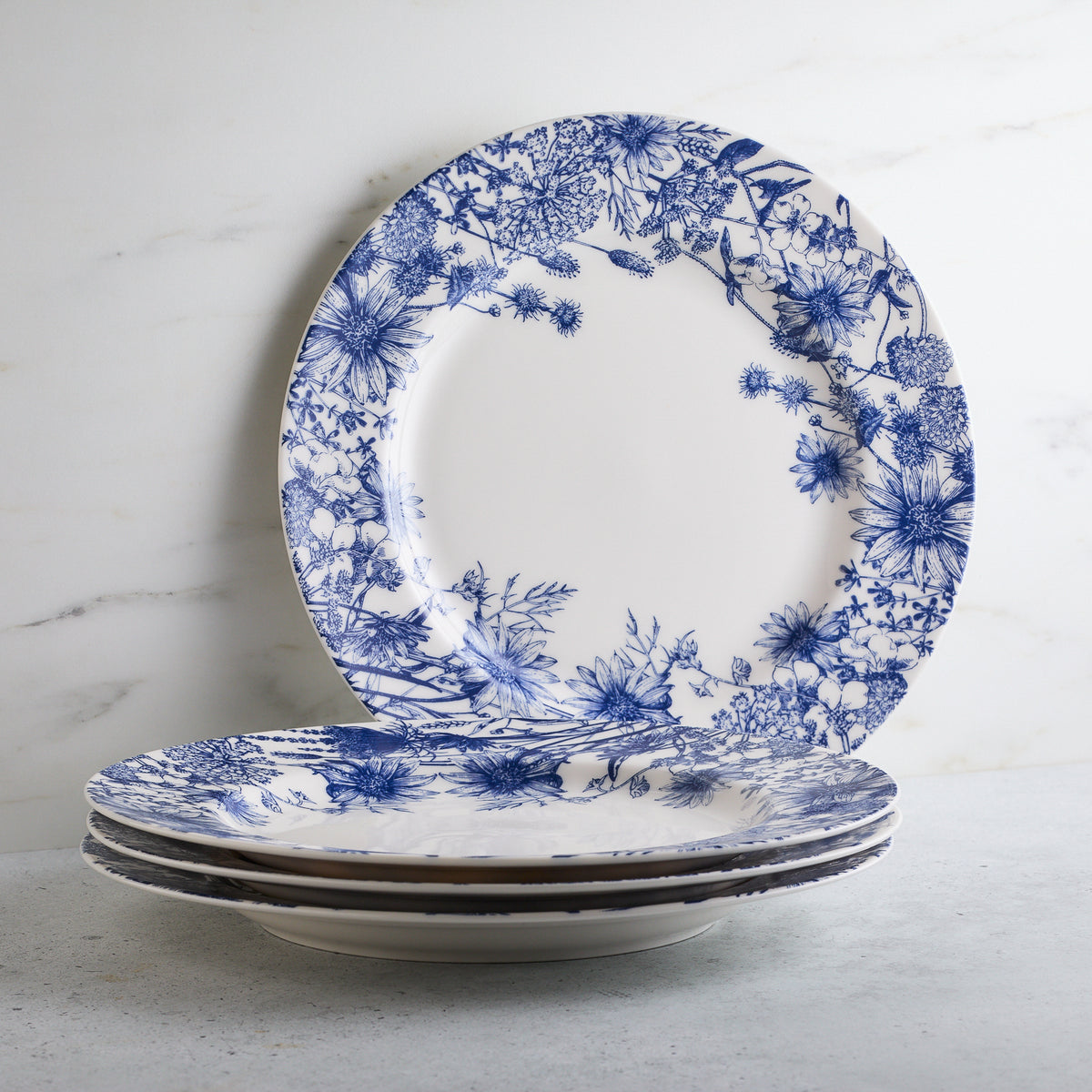 A stack of Caskata Artisanal Home Summer Blues Rimmed Dinner Plates, crafted from high-fired porcelain, set against a light-colored, possibly marble, background.