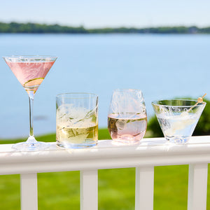 Four drinks in various glassware, including Caskata Artisanal Home Lucy Stemless Martini Glasses and part of a crystal glassware collection, with ice cubes are placed on a white railing overlooking a body of water and greenery in the background.