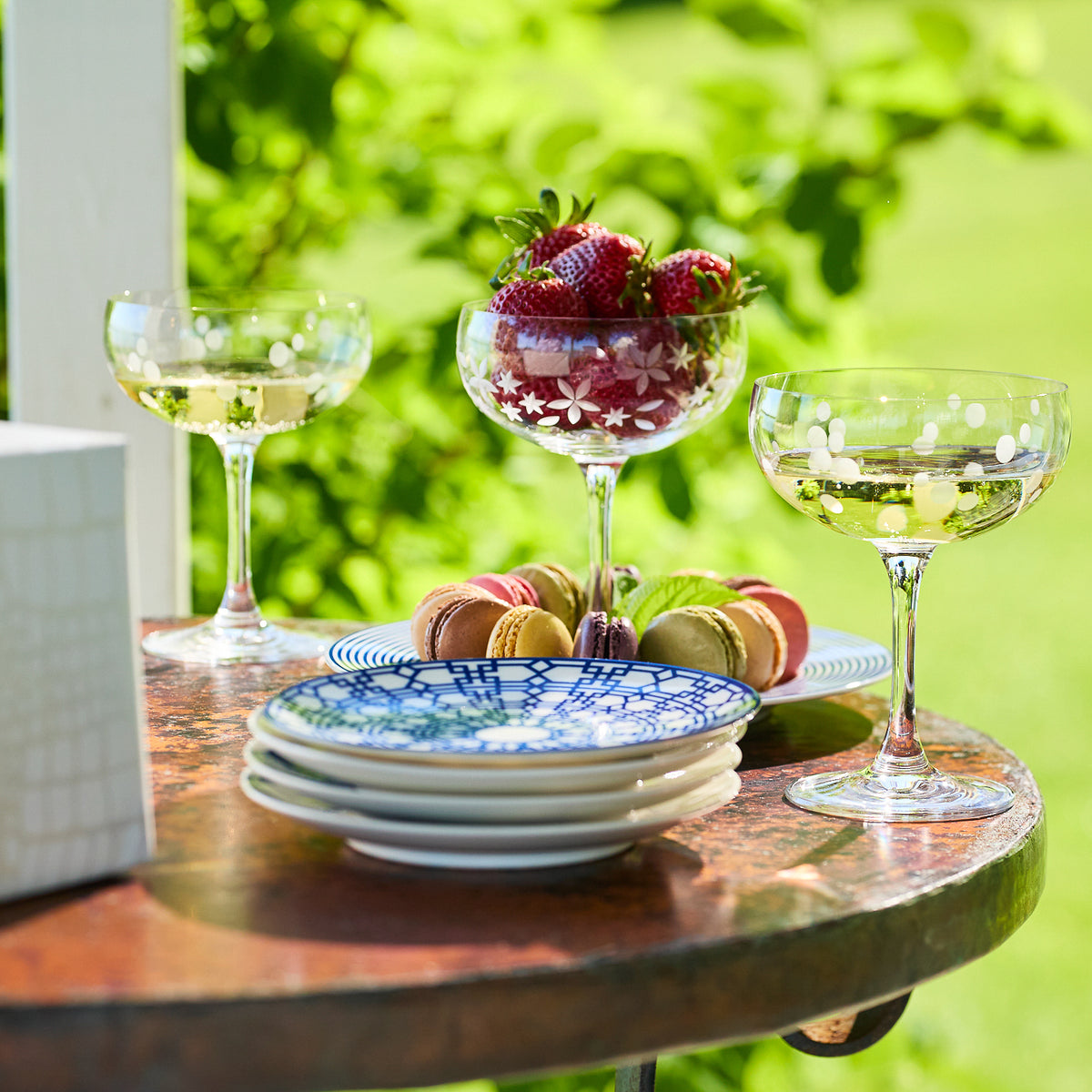 A table set with two glasses of lead-free Chatham Pop Coupe Cocktail Glasses by Caskata, a dessert glass filled with strawberries, a stack of blue and white plates from the Chatham Collection, and colorful macarons in an outdoor setting with lush greenery.