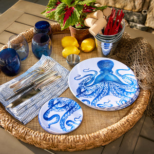 A wicker tray holds Caskata's Lucy Melamine Dinner Plates, Set of 4, featuring a blue octopus theme. Included are BPA-free plates, blue glasses, lemons, metal cutlery, a plant, and a metal container with red utensils. A fishing net drapes on the side for a seaside vibe.