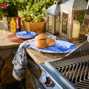 The outdoor kitchen setup showcases sesame buns on a BPA-free Lucy Melamine Oval Rimmed Platter by Caskata, surrounded by condiments. Nearby, a wooden box with artichokes and lanterns sits on the stone countertop beside an elegant grill, all featuring unbreakable style.