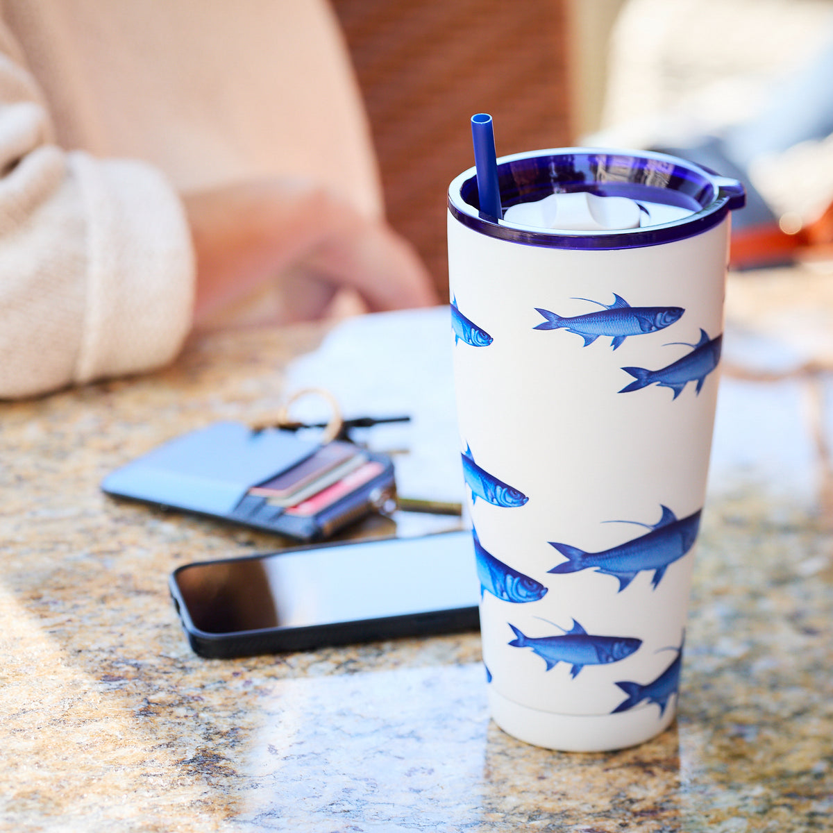 The School of Fish Stainless Steel Insulated Tumbler by Caskata, featuring a blue fish design and a straw, sits on the table next to a smartphone and a wallet with visible cards. This stylish piece of drinkware is perfect for any setting.