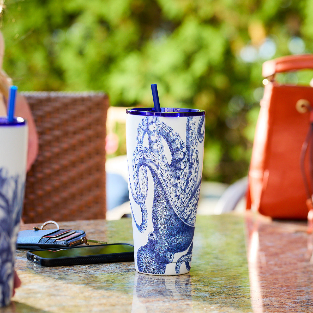 A Lucy Stainless Steel Insulated Tumbler by Caskata, featuring a blue and white octopus design, sits on a granite table. Nearby are a smartphone, keys, and a blurred red bag.