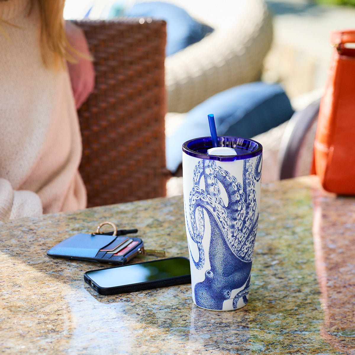 A Lucy Stainless Steel Insulated Tumbler by Caskata with an octopus design, a smartphone, and a wallet with keys are placed on a marble table. A person in a white sweater is partially visible in the background.