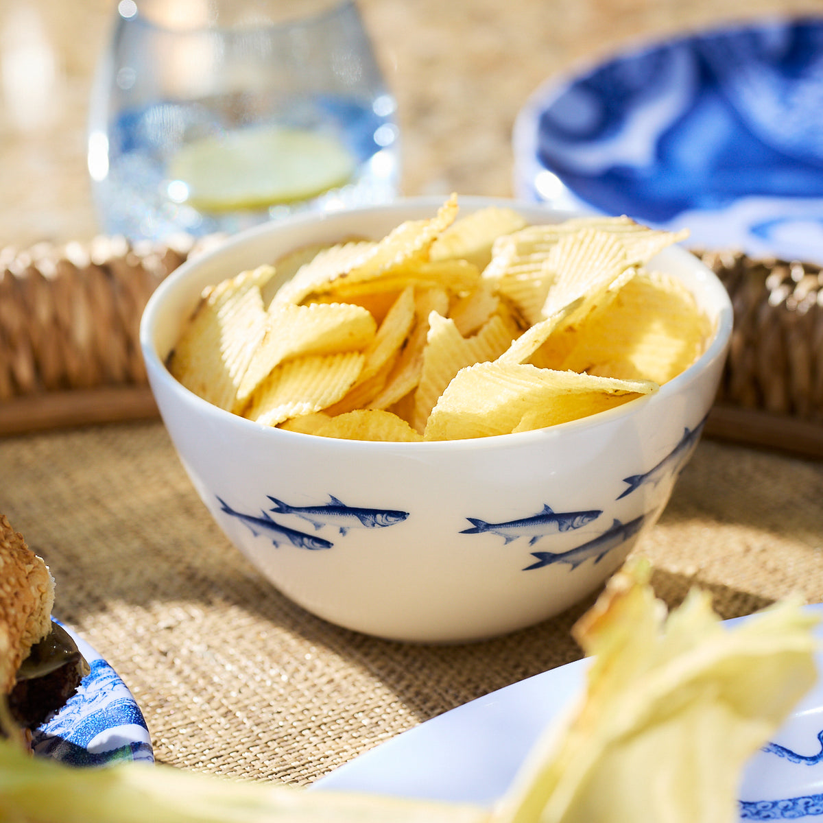 A Caskata School of Fish Cereal Bowl, adorned with a school of fish design, is filled with ridged potato chips, placed on a woven placemat next to plates and a glass of water with lemon.