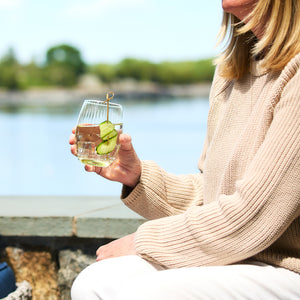 A person wearing a beige sweater holds a glass of water with cucumber slices in a Quinn Citrine Tumbler by Caskata Artisanal Home, sitting by a body of water.