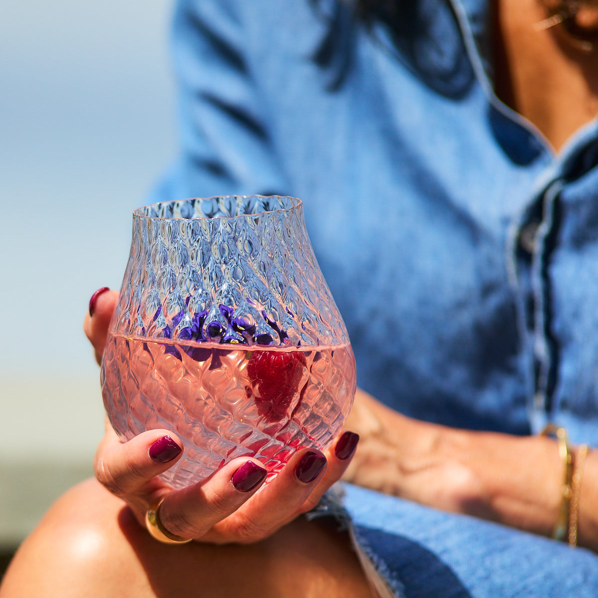A person wearing a denim shirt holds a Caskata Phoebe Clear Stemless Wine Glasses containing a pink beverage with flower petals.