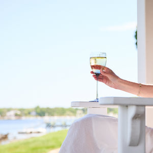 A person holding a glass of white wine in Quinn Ocean White Wine Glasses from Caskata Artisanal Home while sitting at a table outdoors with a scenic view of water and greenery in the background.