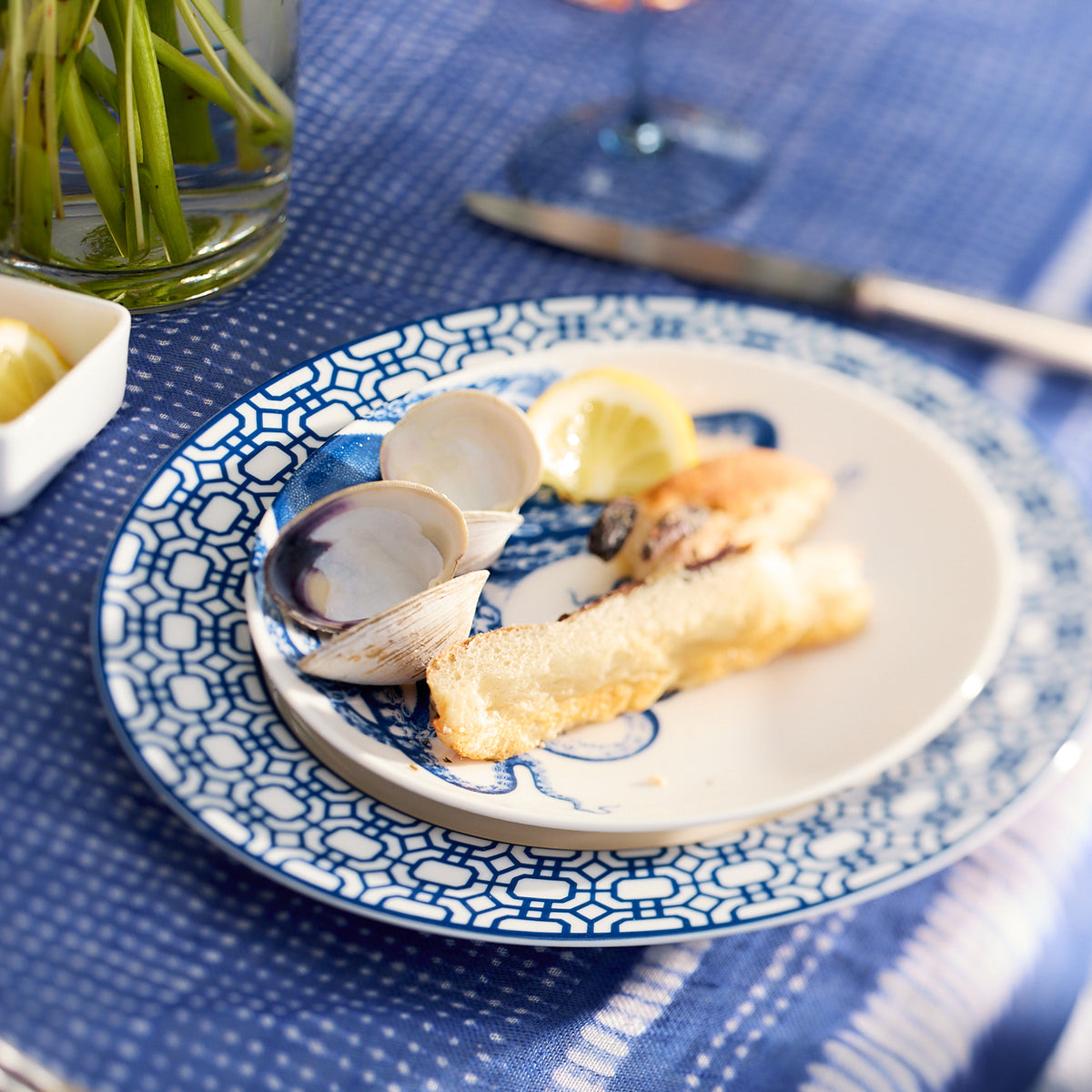 A plate with fried fish sticks, clams in shells, and lemon wedges sits elegantly on a blue and white patterned tablecloth, adding a contemporary note. The dish is presented on the Newport Garden Gate Rimmed Salad Plate from Caskata Artisanal Home.