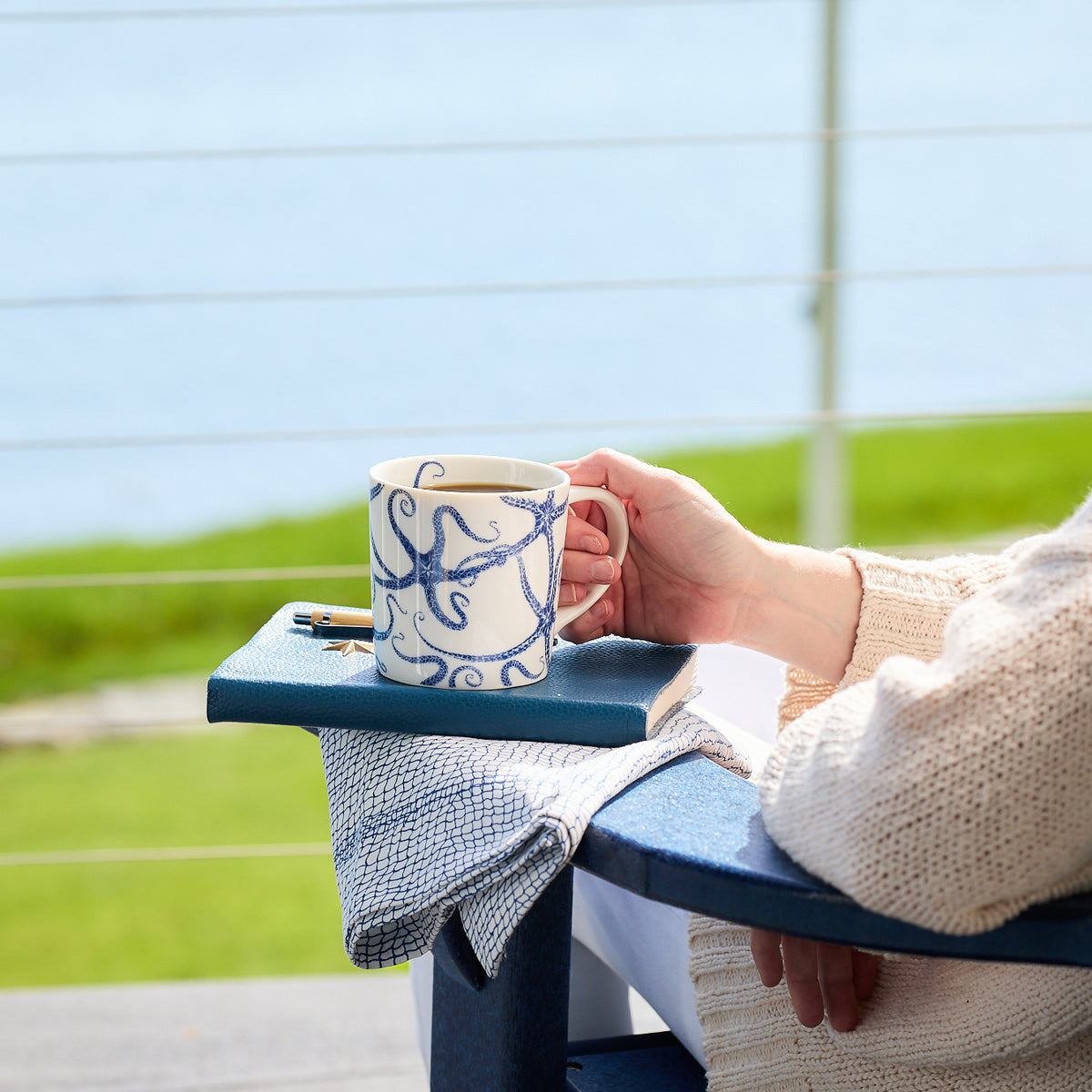 A person holds a high-fired porcelain mug with an octopus design, resting on a chair outside with a body of water and greenery in the background. A notebook and pen sit on the armrest. The Starfish Mug by Caskata Artisanal Home is dishwasher and microwave safe, perfect for enjoying coffee amid nature&#39;s tranquility.