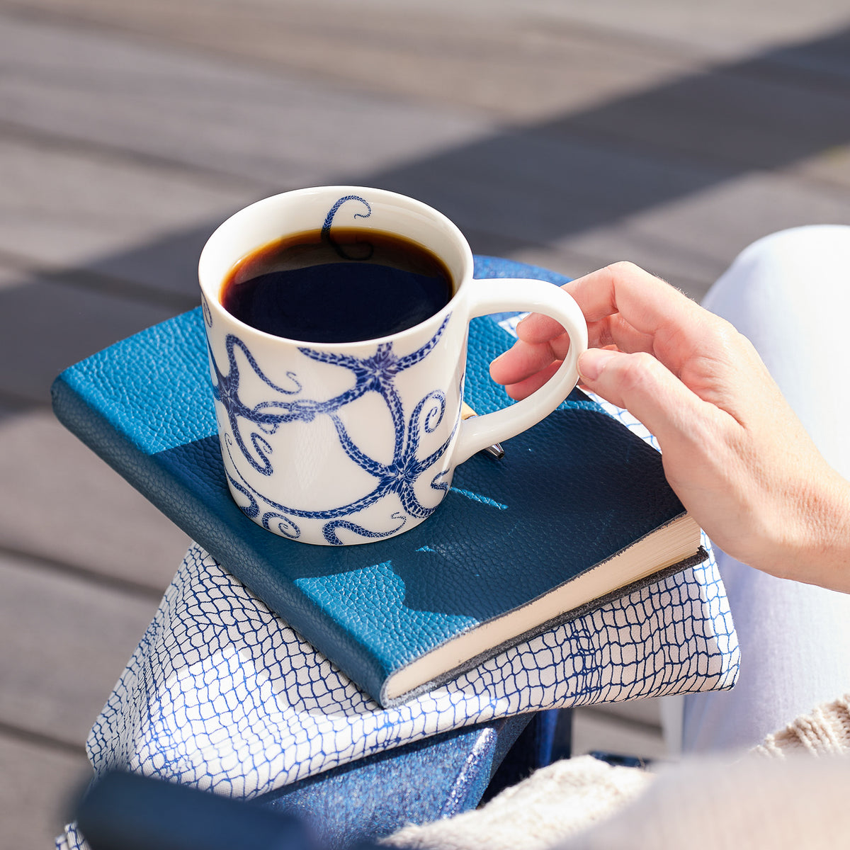 A person holding a Caskata Artisanal Home Starfish Mug filled with coffee, placed on top of two closed blue notebooks. The sturdy and stylish dishwasher-safe mug adds a touch of coastal charm to any moment.