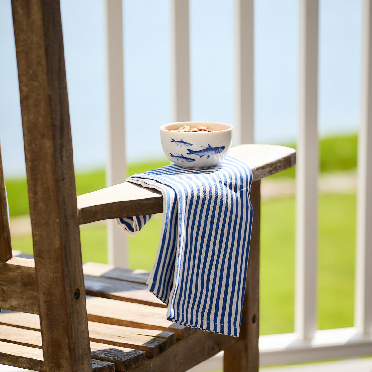 A wooden chair showcases Caskata&#39;s Pinstripe Dinner Napkins in Blue draped over the armrest. A blue fish design bowl holds cereal, while blurred greenery and a railing frame the serene scene in the background.