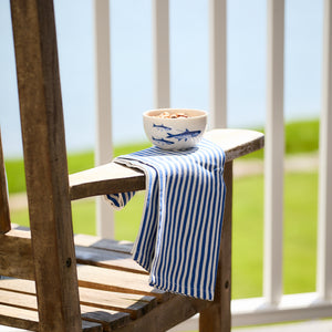 A wooden chair showcases Caskata's Pinstripe Dinner Napkins in Blue draped over the armrest. A blue fish design bowl holds cereal, while blurred greenery and a railing frame the serene scene in the background.