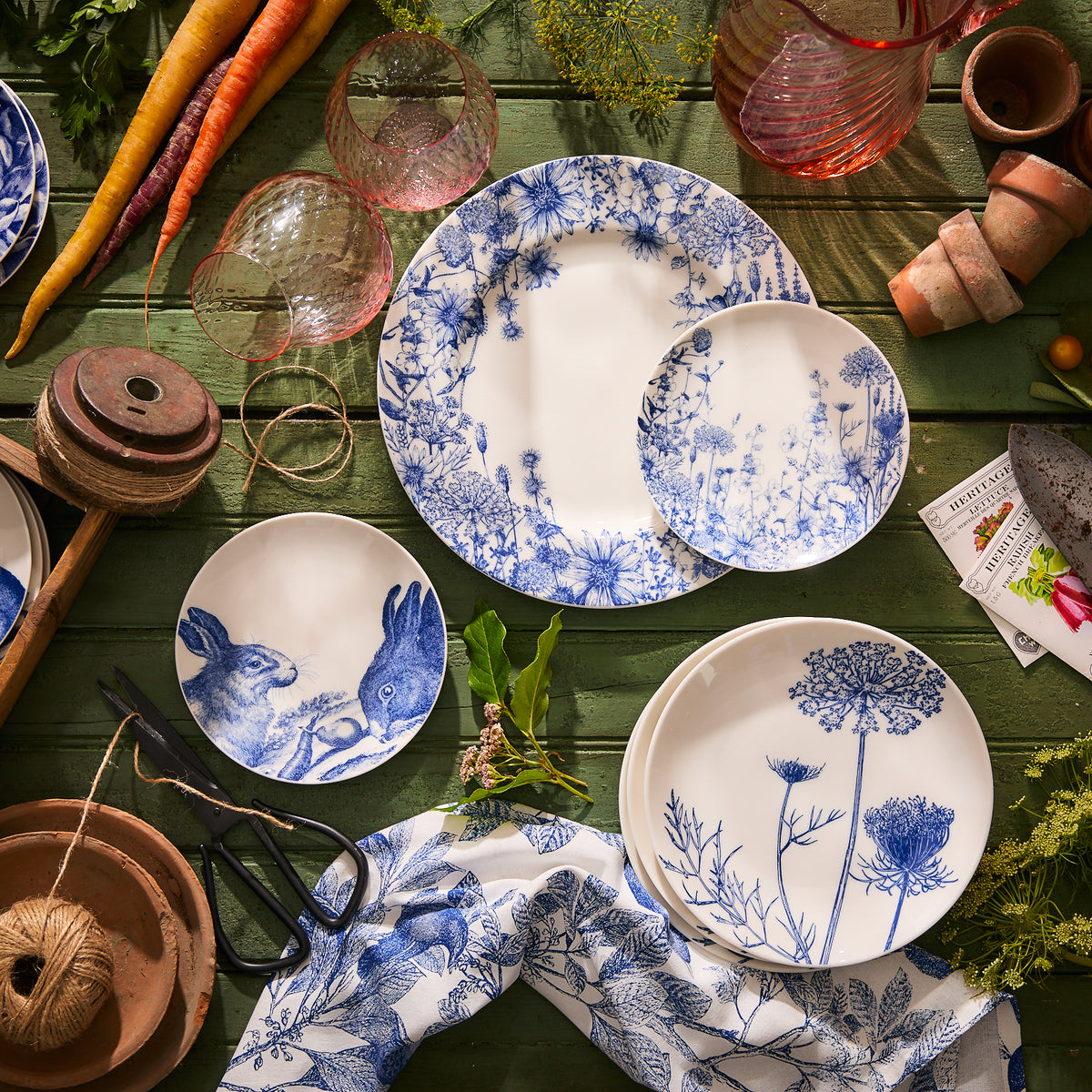 A collection of Caskata&#39;s Summer Blues Small Plates featuring floral and wildlife themes are displayed on a green wooden surface, surrounded by gardening tools, glassware, and vegetables.